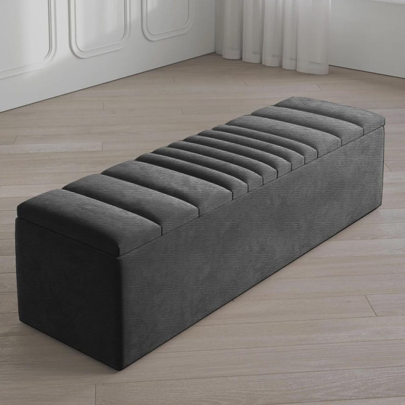 May include: A dark gray upholstered storage bench with a rectangular shape. The top features a series of parallel, padded sections. The bench is placed on a light-colored wooden floor, and the background includes a white wall and a window.