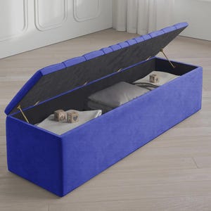 May include: A blue storage ottoman with a hinged lid, revealing interior storage. The ottoman is rectangular and upholstered in a vibrant blue fabric. Inside, there are folded blankets and pillows. Wooden blocks with letters are visible.