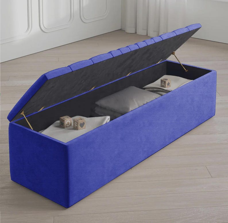 May include: A blue storage ottoman with a hinged lid, revealing interior storage. The ottoman is rectangular and upholstered in a vibrant blue fabric. Inside, there are folded blankets and pillows. Wooden blocks with letters are visible.