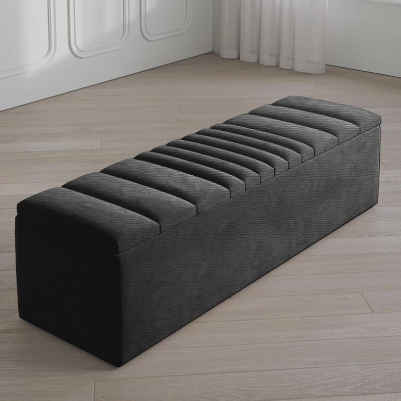 May include: A dark gray upholstered storage bench with a rectangular shape. The bench features a series of vertical, padded sections across the top. The bench is placed on a light-colored wooden floor.