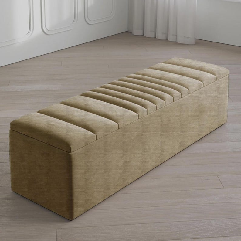 May include: A rectangular, beige storage bench with a ribbed, padded top. The bench is made of a textured fabric and sits on a light-colored wooden floor. The bench is a neutral color and would fit in many home decor styles.
