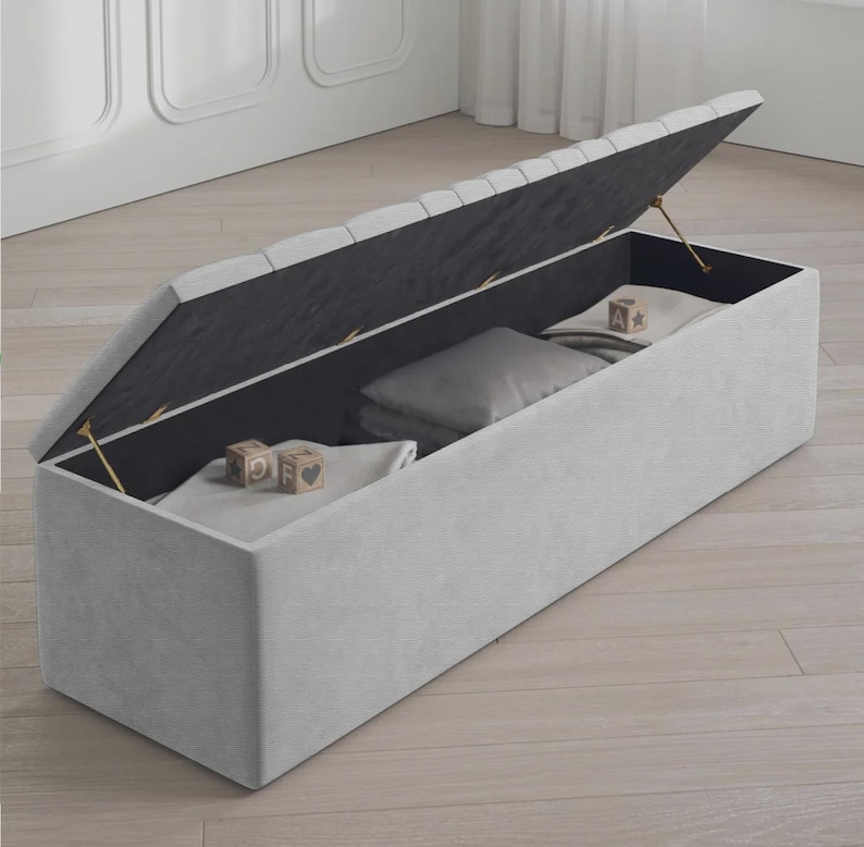 May include: A light gray rectangular storage bench with a hinged lid, partially open. The interior is black, and the bench contains pillows and folded blankets. Wooden blocks with letters and symbols are visible on top of the folded blankets.