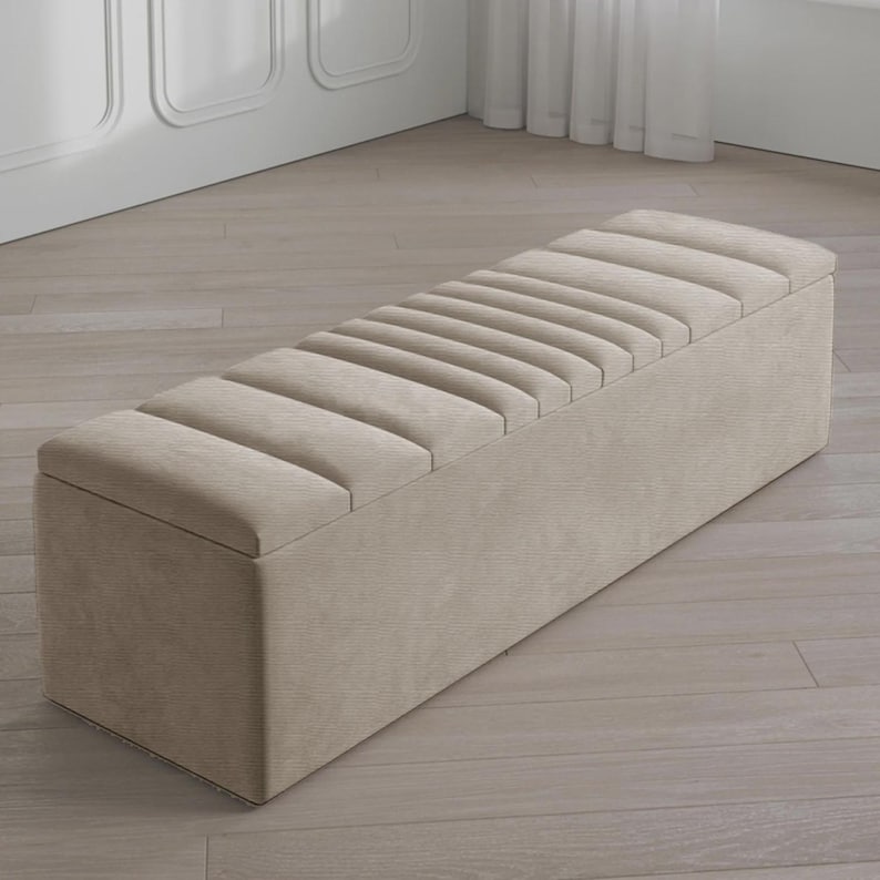 May include: A beige rectangular storage bench with a ribbed, padded top. The bench is made of a textured fabric and has a hinged lid for storage. The neutral color and simple design make it suitable for various interior styles.