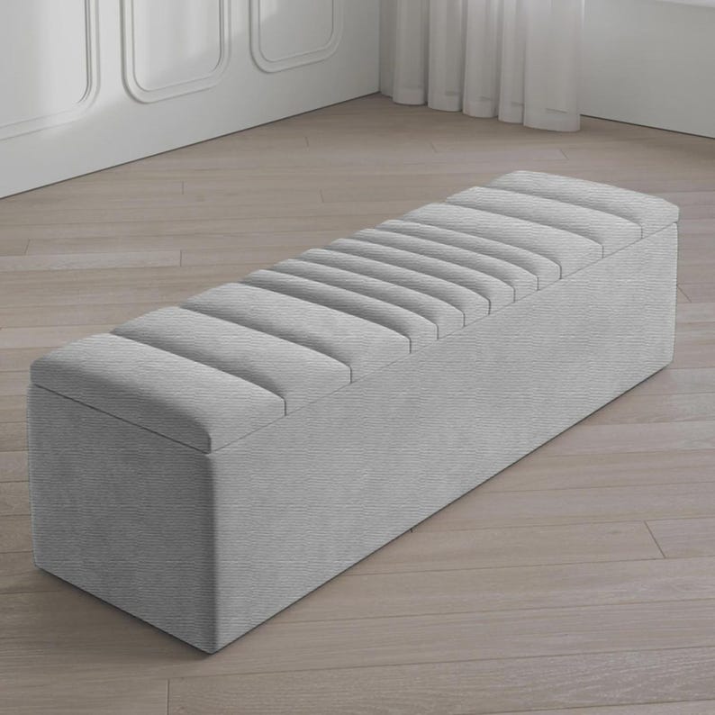 May include: A rectangular, light gray storage bench with a padded, ribbed top. The bench is upholstered in a textured fabric and has a clean, modern design. The bench is placed on a light wood floor.