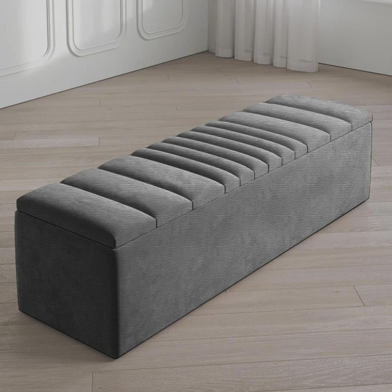 May include: A gray upholstered storage bench with a rectangular shape. The top features a series of parallel, padded sections. The bench is made of a soft, textured fabric and is placed on a light-colored wooden floor.