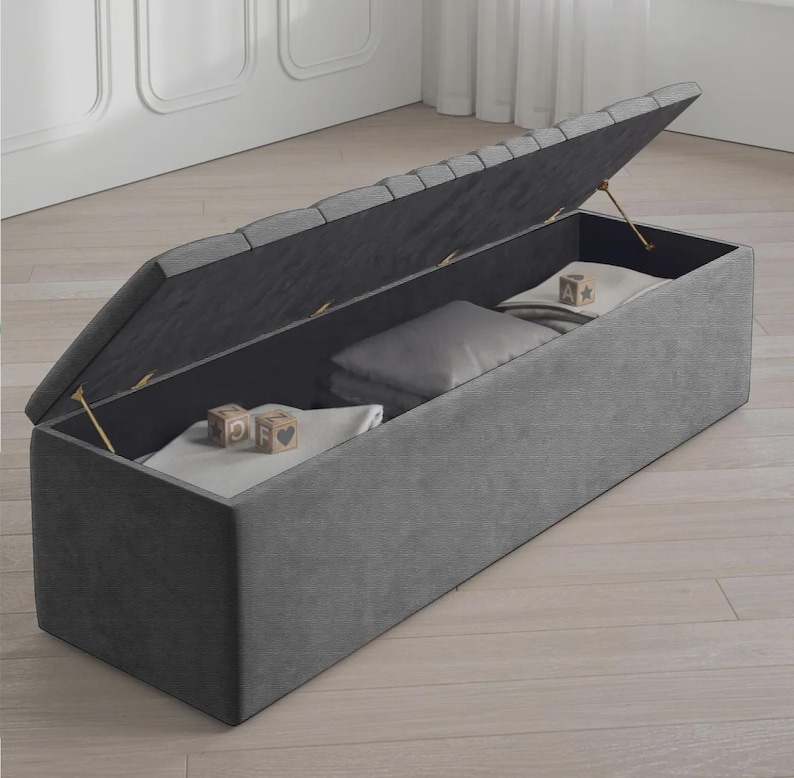 May include: A gray upholstered storage bench with the lid partially open, revealing folded blankets, a pillow, and wooden blocks. The bench is rectangular and has a tufted lid. The interior is dark gray.