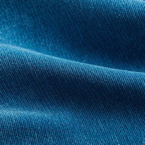 Op de afbeelding: Close-up van een getextureerde, diepblauwe corduroy stof. De stof heeft een geribbeld patroon en een zacht, fluwelig uiterlijk. De plooien en schaduwen creëren diepte en benadrukken de textuur van het materiaal. De kleur is een rijk, verzadigd blauw.