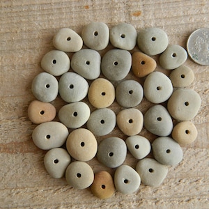 May include: A collection of smooth, grey and tan drilled stones arranged on a wooden surface. A coin is placed near the stones for size comparison. The stones vary in size and shape, with a hole drilled through the centre of each.