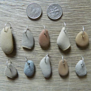 Peut inclure: Une collection de dix petits pendentifs en pierre lisse de différentes formes et couleurs, chacun avec une boucle argentée pour accrocher. Deux pièces de monnaie sont placées en haut. Les pendentifs sont exposés sur une surface en bois.
