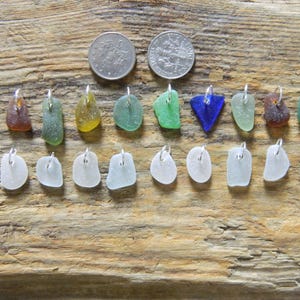 Könnte beinhalten: Eine Sammlung von zwanzig Meerglas-Anhängern mit silberfarbenen Ösen, angeordnet in zwei Reihen. Die obere Reihe zeigt verschiedene Farben wie Blau, Braun, Grün, Gelb und Transparent. Die untere Reihe besteht aus zehn weißen Meerglas-Anhängern.