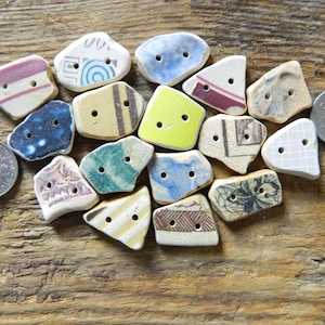 Pode incluir: Uma coleção de botões de cerâmica de formato irregular em várias cores e padrões, incluindo azul, amarelo e bege. Cada botão tem dois furos para costura. Uma moeda é incluída para referência de tamanho. Os botões estão dispostos sobre uma superfície de madeira.