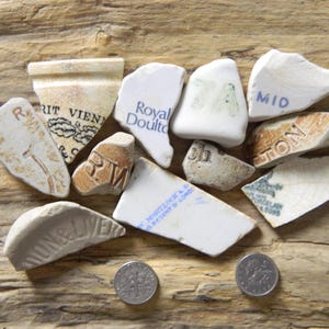 Può includere: Una collezione di undici frammenti di ceramica bianca con vari testi e loghi stampati in blu e nero, inclusi frammenti contrassegnati Royal Doulton e altre parole parziali, accompagnati da due monete color argento per riferimento di scala.
