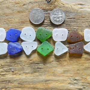 May include: A collection of fourteen irregular, frosted sea glass buttons arranged in two rows. Each button features two small drilled holes. The colours include white, cobalt blue, green, and brown. Two silver coins are placed above the buttons.