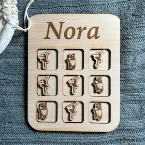 May include: A wooden board with the name "Nora" engraved on it. The board has nine squares, each with a cutout for a small wooden piece. The pieces are either a ballerina or a pair of ballet shoes.
