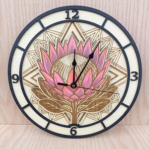 Könnte beinhalten: Eine runde Wanduhr mit geschichtetem Holzdesign und einer zentralen Illustration einer rosa-braunen Protea-Blüte. Das Zifferblatt hat einen hellen Holzhintergrund mit geometrischem Muster, schwarzen Ziffern 3, 6, 9, 12 und schwarzen Zeigern.