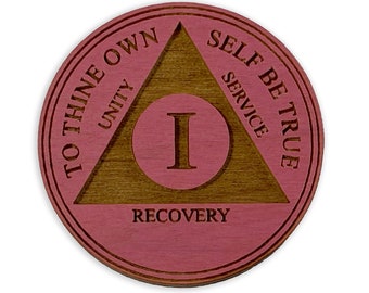 Puede incluir: Una ficha de sobriedad redonda de madera con un acabado rosa y marrón. La ficha presenta un triángulo con las palabras "To Thine Own Self Be True" y las palabras "Unity, Service, Recovery" inscritas en ella. El número "I" está en el centro del triángulo.