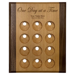 May include: A wooden plaque with a walnut border and a light wood center. The plaque has 12 circular cutouts and the text "One Day at a Time" engraved in a script font. The text "Your Name Here" and "Recovery Date Here" are also engraved on the plaque.