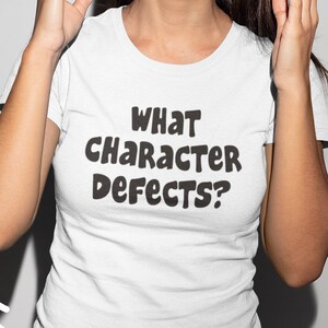 Peut inclure: Une femme portant des lunettes de soleil roses et un t-shirt blanc avec le texte "What Character Defects?" imprimé dessus. Elle sourit et lève les mains en l'air. L'arrière-plan est de couleur gris clair.