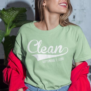 May include: A light green t-shirt with a white graphic that says "Clean" with a baseball-style font and the date "September 7, 1998" underneath. The model is wearing the shirt with a red jacket and blue and white striped jeans.