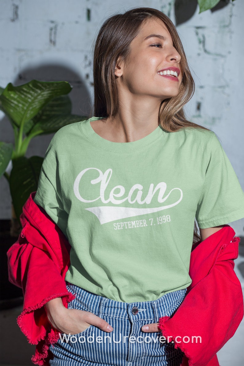 May include: A light green t-shirt with a white graphic that says "Clean" with a baseball-style font and the date "September 7, 1998" underneath. The model is wearing the shirt with a red jacket and blue and white striped jeans.