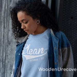 May include: A light blue sweatshirt with the word "Clean" in white script with a white baseball-style arc underneath. The text "SEPTEMBER 7, 1990" is printed below the word "Clean". The sweatshirt is being worn by a person with curly black hair and a denim jacket.