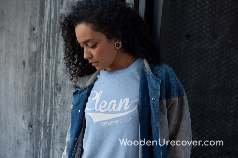 May include: A light blue sweatshirt with the word "Clean" in white script with a white baseball-style arc underneath. The text "SEPTEMBER 7, 1990" is printed below the word "Clean". The sweatshirt is being worn by a person with curly black hair and a denim jacket.