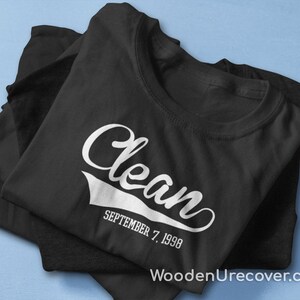 May include: Black t-shirt with white text that reads "Clean" in a vintage style font with a white baseball-style arc underneath. The text "SEPTEMBER 7, 1998" is below the arc.