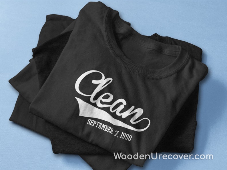 May include: Black t-shirt with white text that reads "Clean" in a vintage style font with a white baseball-style arc underneath. The text "SEPTEMBER 7, 1998" is below the arc.