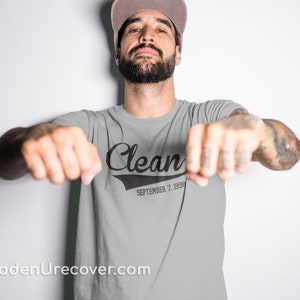 May include: A gray t-shirt with a black graphic that says "Clean" with a baseball-style font and "September 7, 1998" underneath. The person wearing the shirt is facing the camera with their arms outstretched.