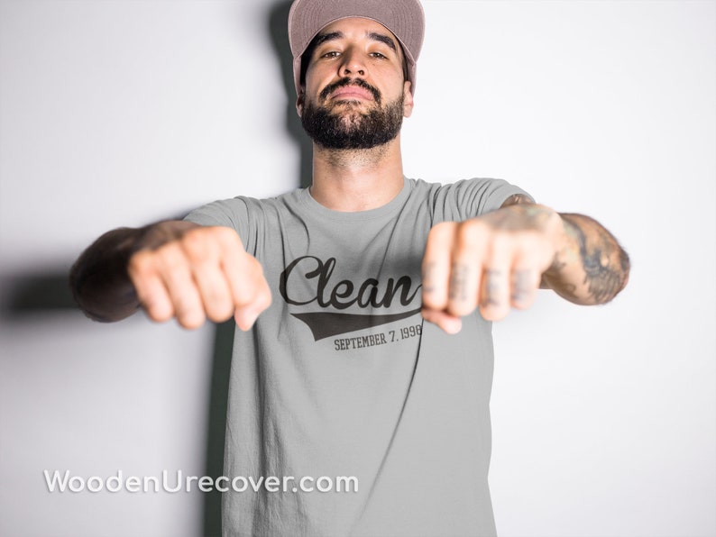 May include: A gray t-shirt with a black graphic that says "Clean" with a baseball-style font and "September 7, 1998" underneath. The person wearing the shirt is facing the camera with their arms outstretched.