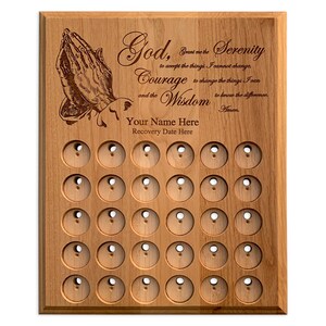 May include: Wooden plaque with engraved praying hands and the Serenity Prayer. Features 30 circular holes for tokens, with space for a name and recovery date. The plaque is made of light brown wood.