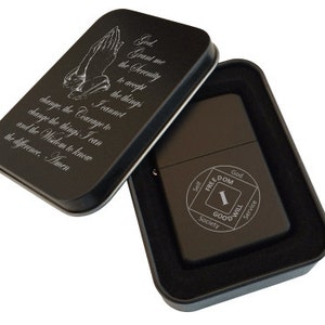 May include: Black lighter with a silver inscription of the Serenity Prayer and a black and silver AA symbol. The lighter is in a black tin box.