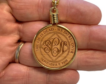 Puede incluir: Collar con colgante dorado y un círculo de madera con la inscripción "The Lie Is Dead - We Do Recover" y un símbolo en forma de diamante con las iniciales "S.C.W." en su interior. La fecha "November 8, 1998" está inscrita debajo del símbolo.