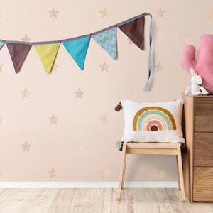 Puede incluir: Escena de habitación infantil con una pared rosa adornada con estrellas doradas. Una guirnalda colorida cuelga arriba, y una almohada de arcoíris descansa sobre una silla de madera. Una almohada en forma de cactus rosa y una figura de conejo blanco están sobre una cómoda de madera.