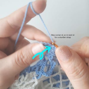 May include: Close-up of hands crocheting with blue and white yarn. The text "Skip corner st, sc in next st for a sturdier strap" is visible. A crochet hook is inserted into the blue yarn, creating a detailed pattern.
