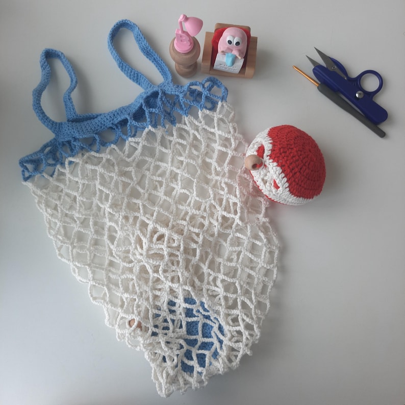 May include: A white crocheted net bag with blue trim and handles. A red and white crocheted ball, small pink lamp, and other small objects are next to the bag. A pair of blue handled scissors and a crochet hook are also present.