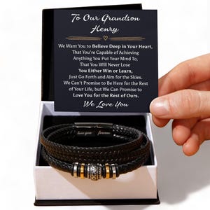 May include: A black leather bracelet with silver and gold details is presented in a gift box. The box includes a card with the text "To Our Grandson Henry" and a loving message. The bracelet features a multi-strand design.