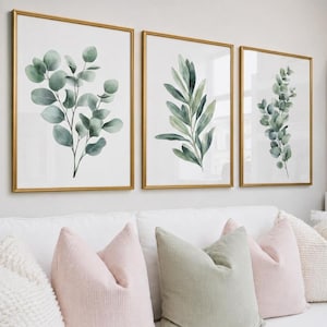 Op de afbeelding: Drie ingelijste botanische prints met gouden frames. Elke print toont een aquarelweergave van groene bladeren en takken op een witte achtergrond. De prints worden weergegeven boven een witte bank met roze en groene kussens.