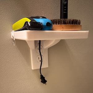 May include: A white shelf with a built-in electrical outlet, holding a blue and yellow electric shaver, a black toothbrush, and a wooden brush. A black power cord hangs below the shelf. The shelf is mounted on a light gray wall.