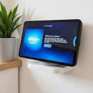May include: A black tablet displaying the Alexa+ welcome screen, mounted on a white shelf. The screen shows the Alexa+ logo and text that reads "Welcome to Alexa+". A small potted plant is visible in the background.