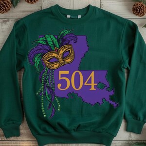 May include: Green sweatshirt featuring a purple outline of Louisiana with the number 504 in gold. A gold and black mask with green and purple feathers and beads is on the left side. The sweatshirt is a solid green color.