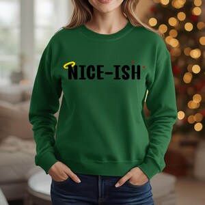 May include: A dark green sweatshirt featuring the black text "NICE-ISH". The word "NICE" has a yellow halo above it, and the "ISH" has two red apostrophes above it. The sweatshirt is a crew neck style.