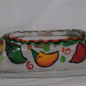 Puede incluir: Un recipiente de mantequilla de vidrio transparente con un diseño pintado a mano de pimientos rojos, amarillos y verdes. El recipiente tiene un borde festoneado y un borde negro y naranja.
