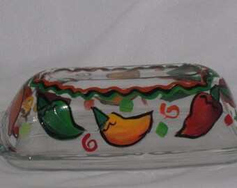 May include: A clear glass butter dish with a hand-painted design of red, yellow, and green peppers. The dish has a scalloped edge and a black and orange border.