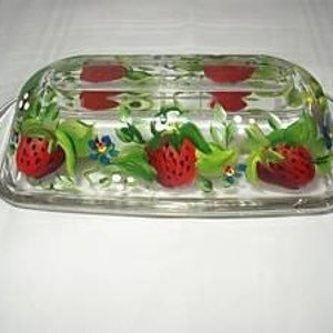 Puede incluir: Un recipiente para mantequilla de vidrio transparente con un diseño pintado a mano de fresas rojas, hojas verdes y flores azules.