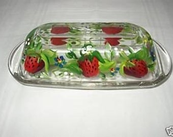 May include: A clear glass butter dish with a hand-painted design of red strawberries, green leaves, and blue flowers.