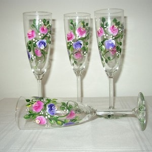 Puede incluir: Cuatro copas de champán con diseños florales pintados a mano. Las copas presentan flores rosas, moradas y verdes sobre un fondo de vidrio transparente.