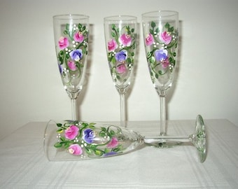 May include: Four champagne flutes with hand-painted floral designs. The glasses feature pink, purple, and green flowers on a clear glass background.
