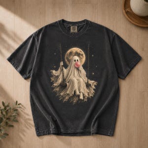 Könnte beinhalten: Ein anthrazitfarbenes Kurzarm-T-Shirt mit einem Grafikdruck eines Geistes, der auf einer Schaukel vor einem großen Vollmond sitzt. Der Geist trägt ein weißes Laken und bläst eine runde rosa Kaugummiblase. Das Design enthält Sterne im Hintergrund.