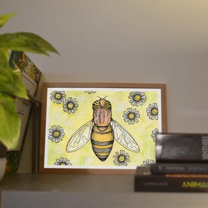 Puede incluir: Una pintura de acuarela enmarcada de una abeja con rayas amarillas y negras, rodeada de margaritas blancas sobre un fondo amarillo. La obra de arte se exhibe en una estantería con libros y una planta en maceta.